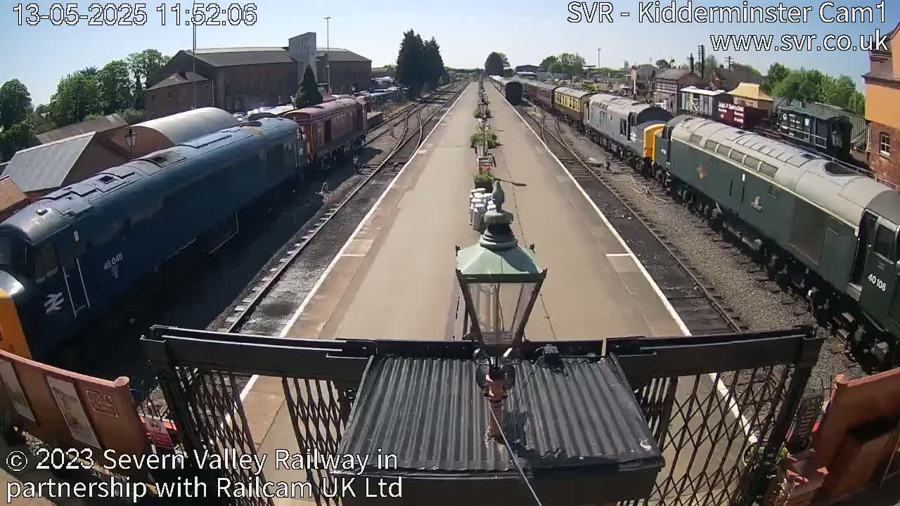 A sunny day at a train station shows multiple locomotives and passenger cars on parallel tracks.