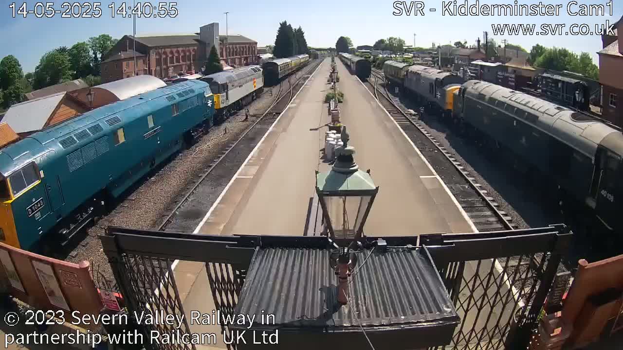 A sunny day at a train station shows multiple locomotives of varying colors and designs parked along parallel tracks.