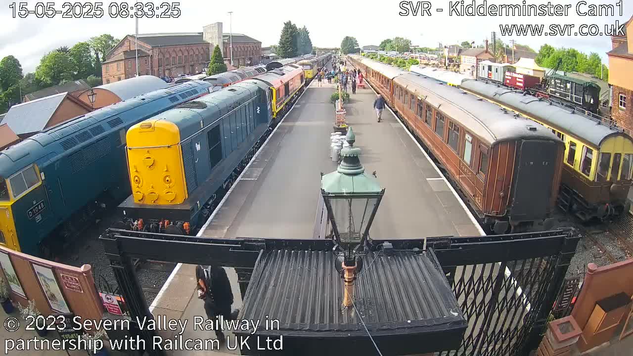 A sunny day at a train station shows numerous locomotives and passenger cars parked on tracks, with a few people walking nearby.