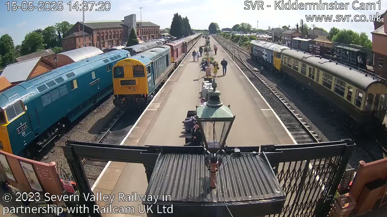 A sunny day at a train station shows several diesel locomotives and passenger cars parked on tracks, with people walking and sitting on the platform.