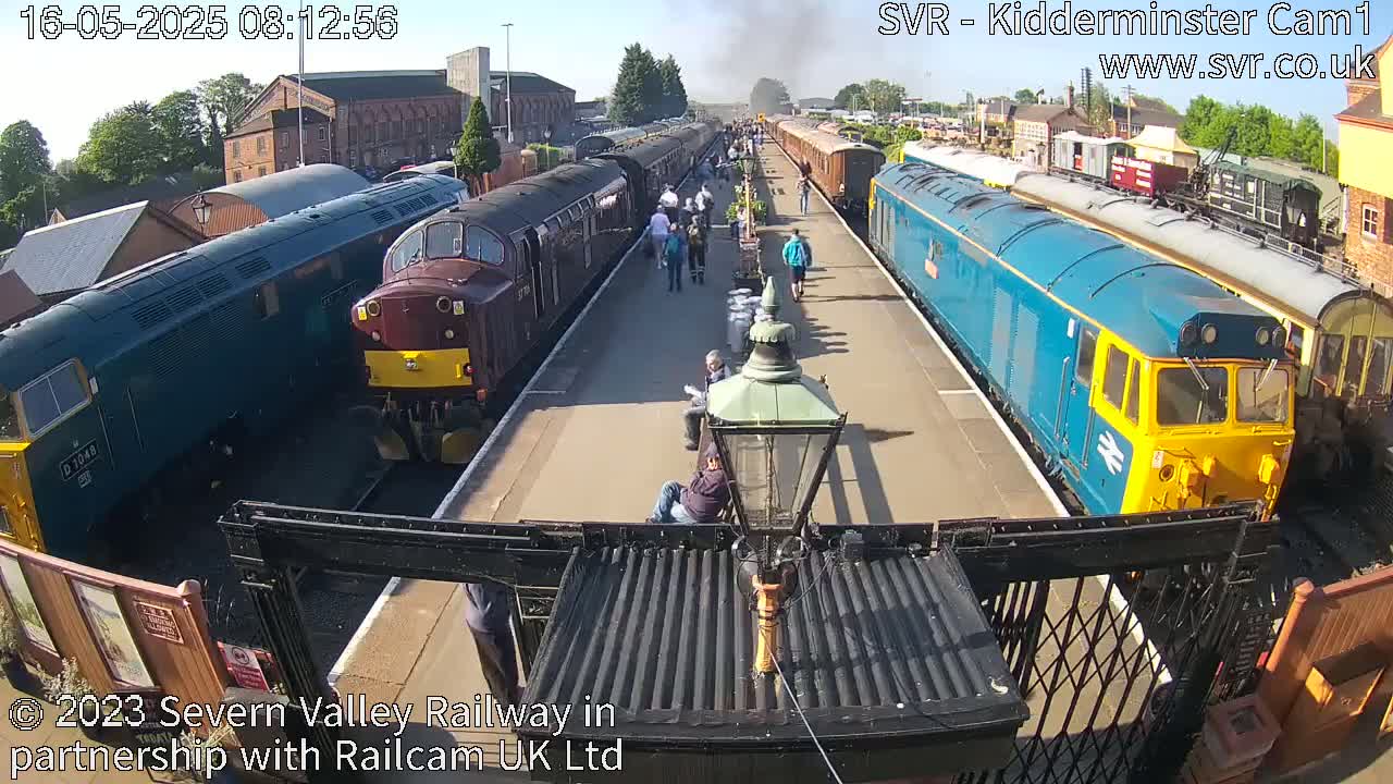 Multiple diesel locomotives and passenger cars are parked at a train station on a sunny day, with several people standing and sitting nearby.