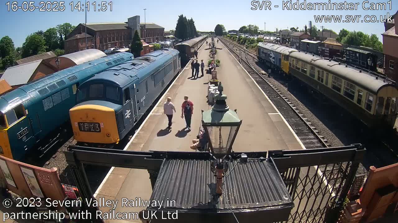 A sunny day at a train station shows several people walking near parked locomotives and railway cars along a platform.