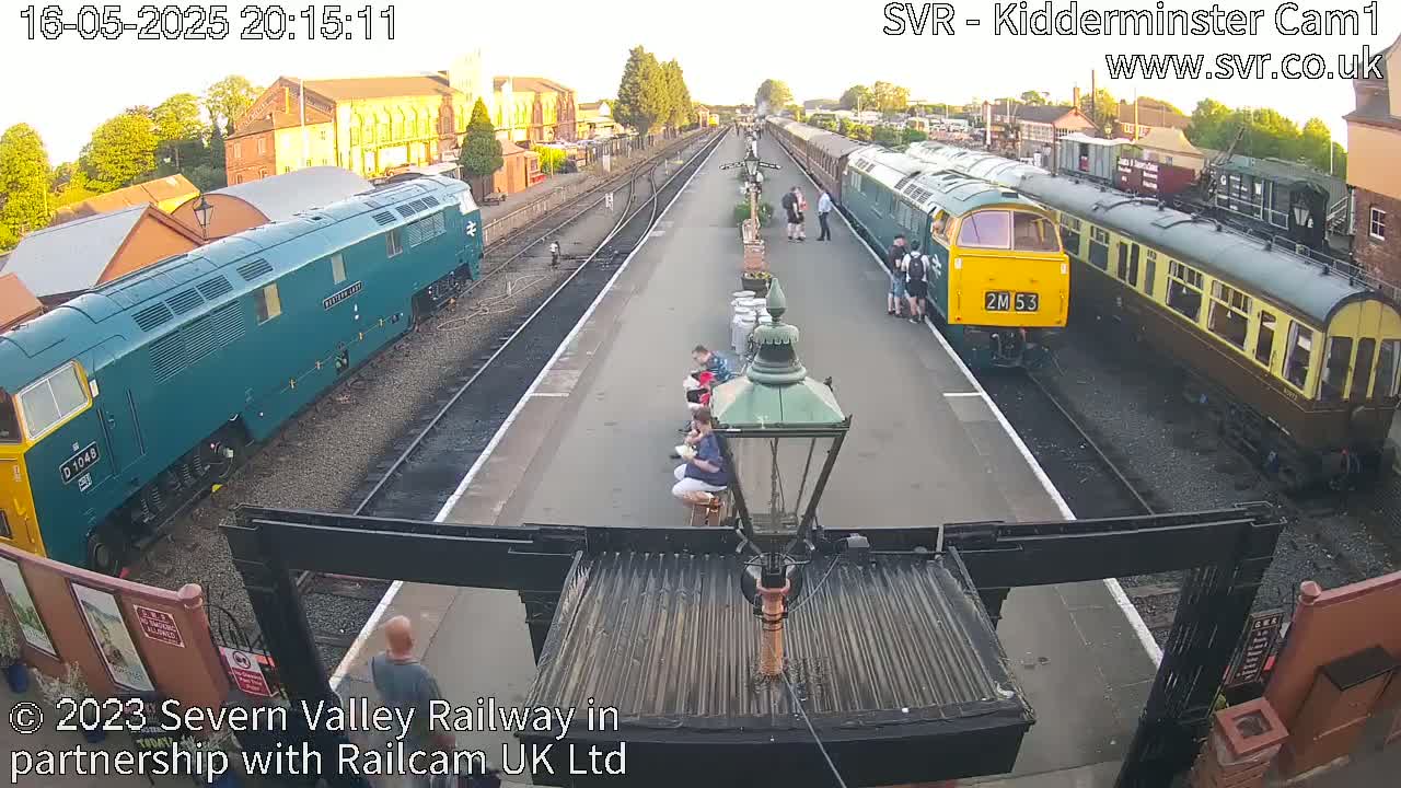 Several teal and green diesel locomotives and passenger cars are parked at a train station at dusk, with several people standing and sitting nearby.