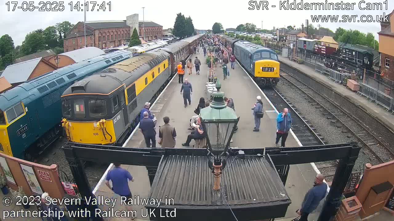 A train station platform with several diesel locomotives and passenger cars, many people milling about, under an overcast sky.
