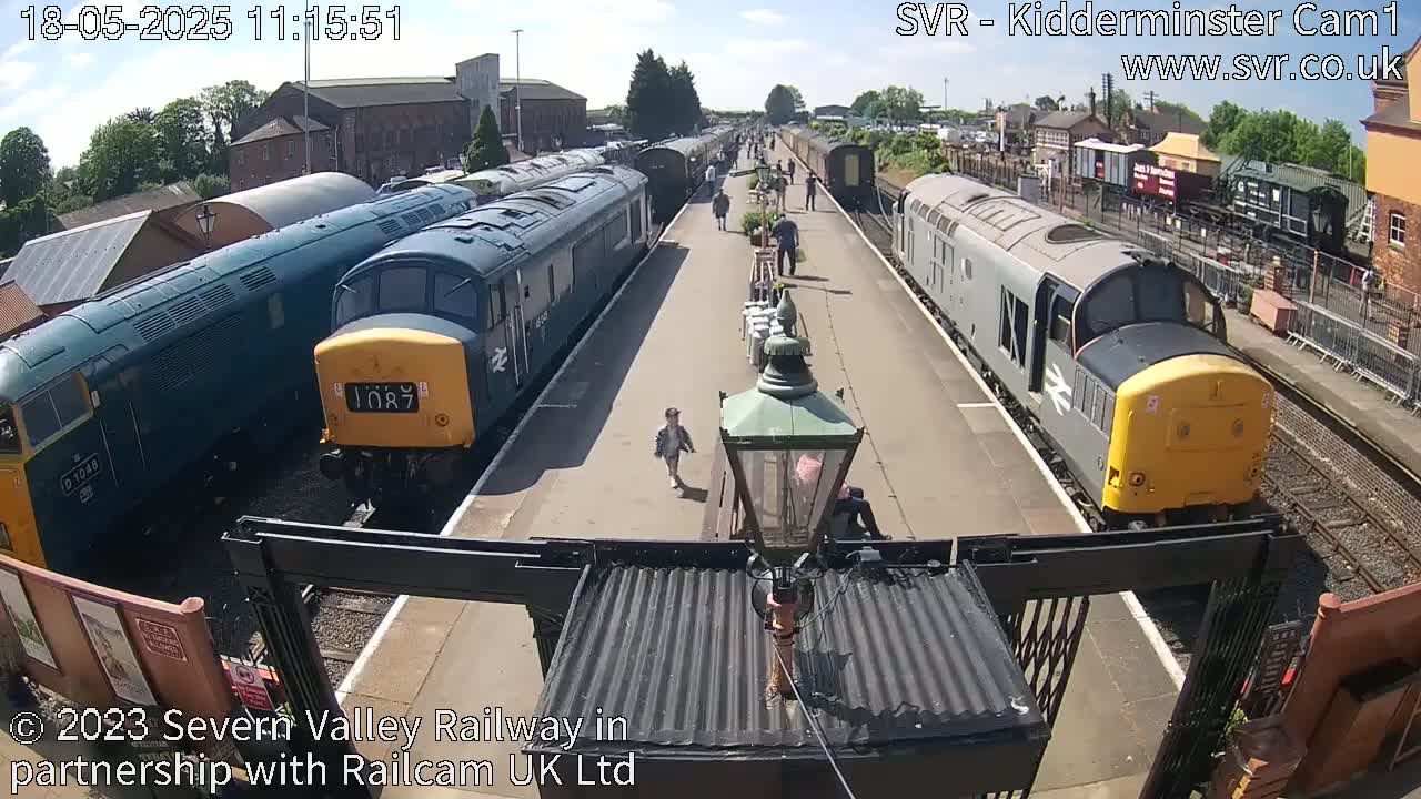 Multiple diesel locomotives are parked at a train station on a sunny day, with a few people walking nearby.