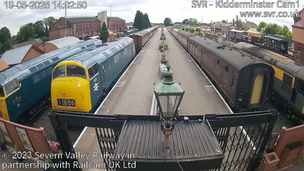 Two teal and yellow diesel locomotives and several passenger cars are parked on a railway platform on an overcast day.