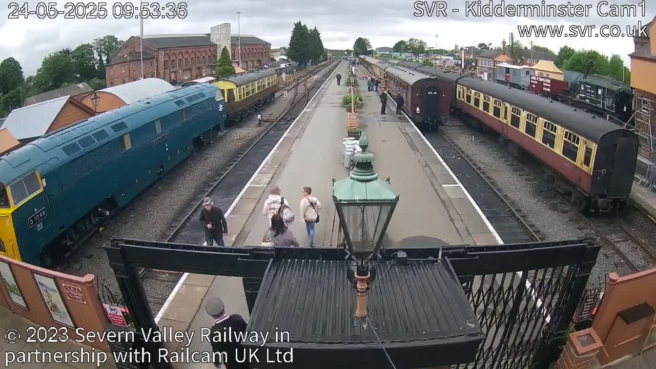 A railway station platform with several train cars and a few people walking on an overcast day.