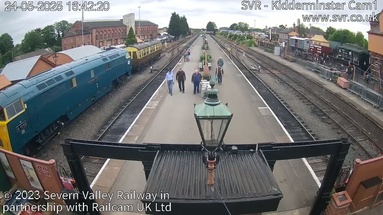 A train station with several people walking along a platform between train tracks on an overcast day.