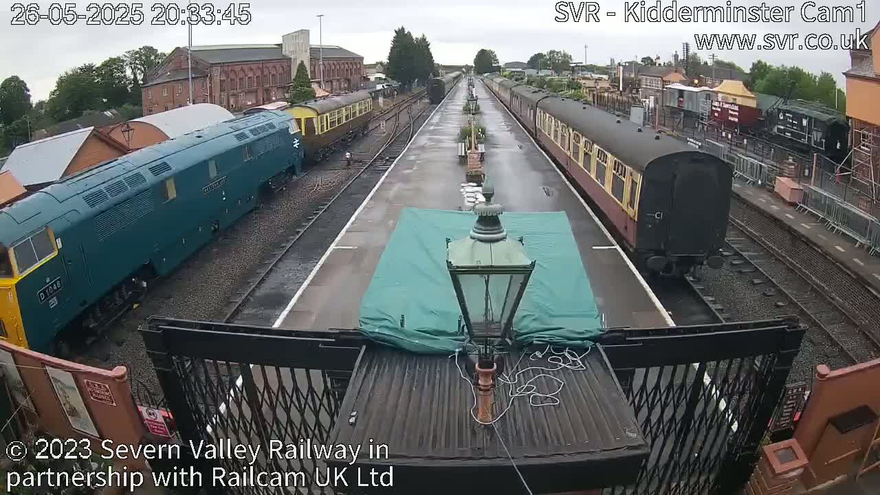 Two passenger trains are visible on a railway platform in the rain.