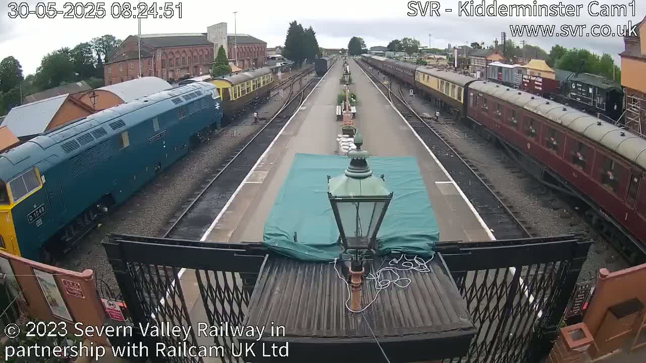 A railway platform with multiple passenger train cars and a diesel locomotive on either side, under an overcast sky.