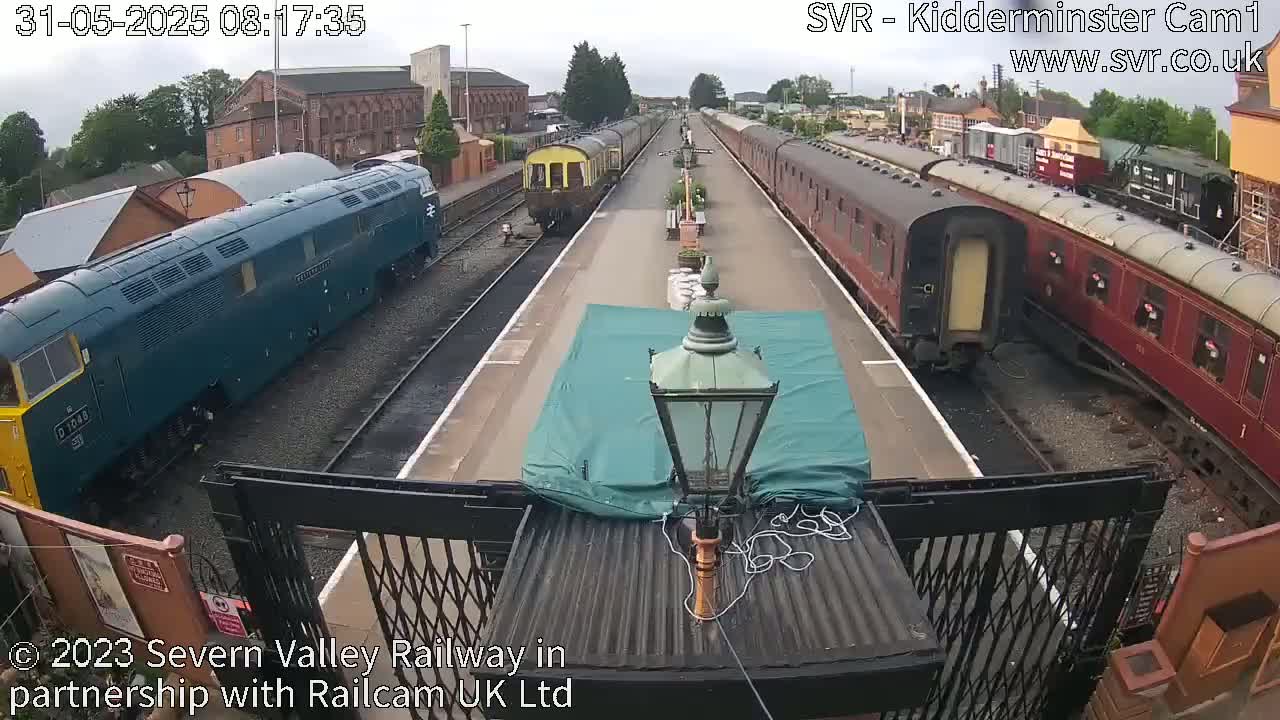 A railway platform with a dark-green diesel locomotive and several passenger cars, under an overcast sky.
