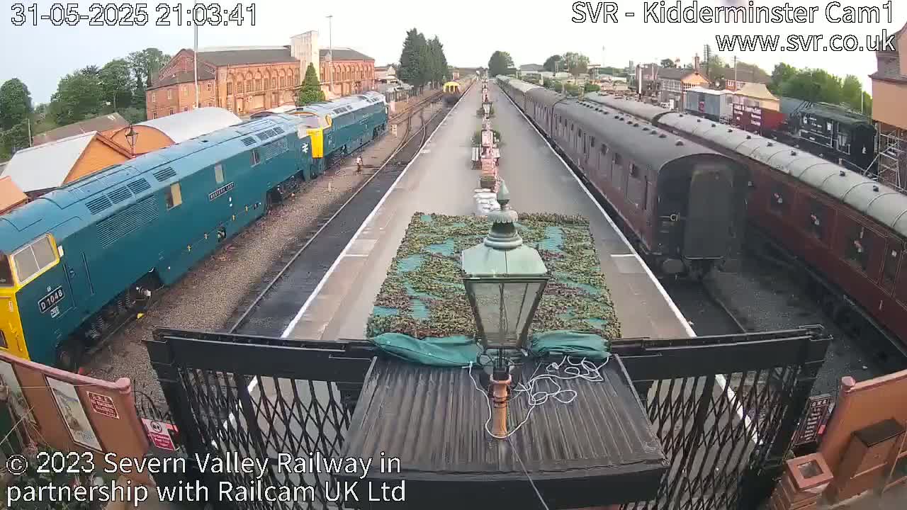 A high-angle view shows two trains, one teal and one maroon, parked on railway tracks under a partly cloudy sky.
