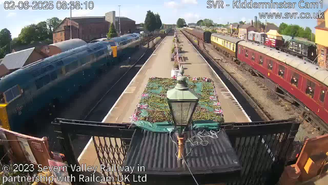 A sunny day at a train station shows two trains, one dark blue and one red, on parallel tracks with a floral display between them.