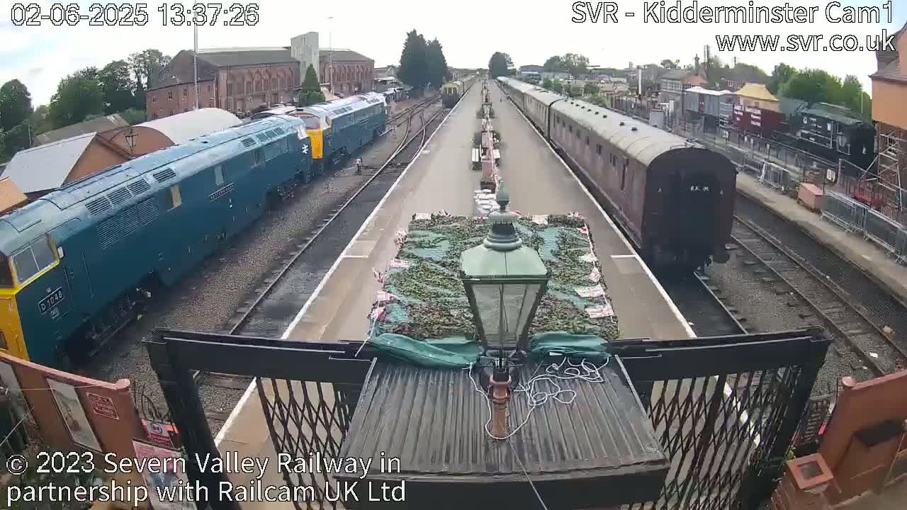 A railway platform with several trains, one teal and one brown, under an overcast sky.
