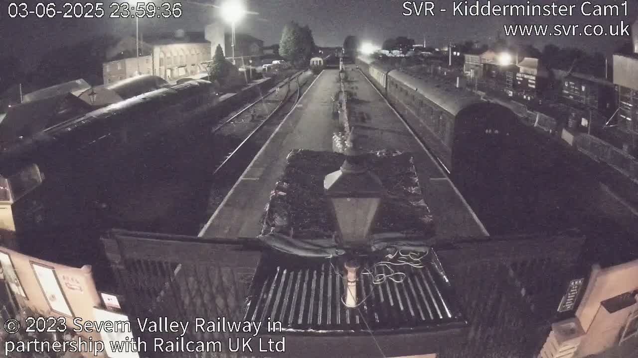 A nighttime view from a low vantage point shows a railway station with several train cars parked on tracks under a dark sky.