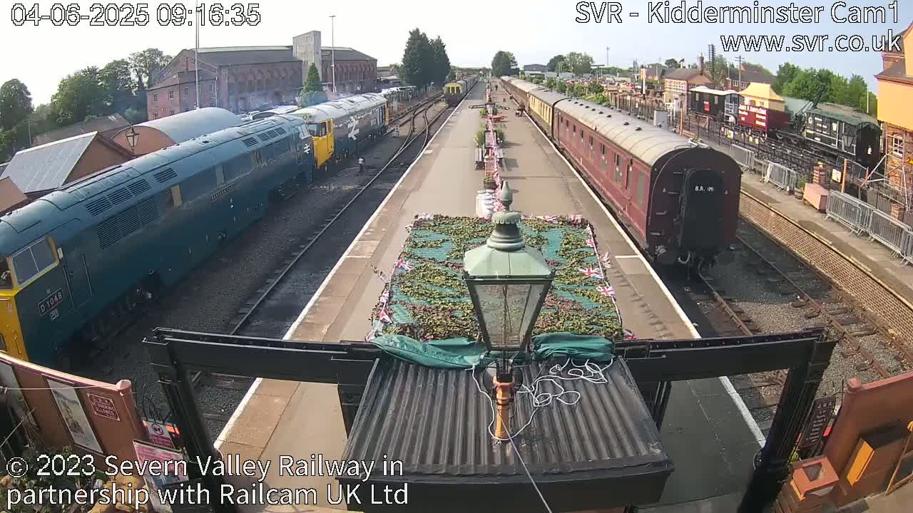 A railway station platform on a sunny day with a dark-blue diesel locomotive and a maroon passenger train at opposite ends of the platform.