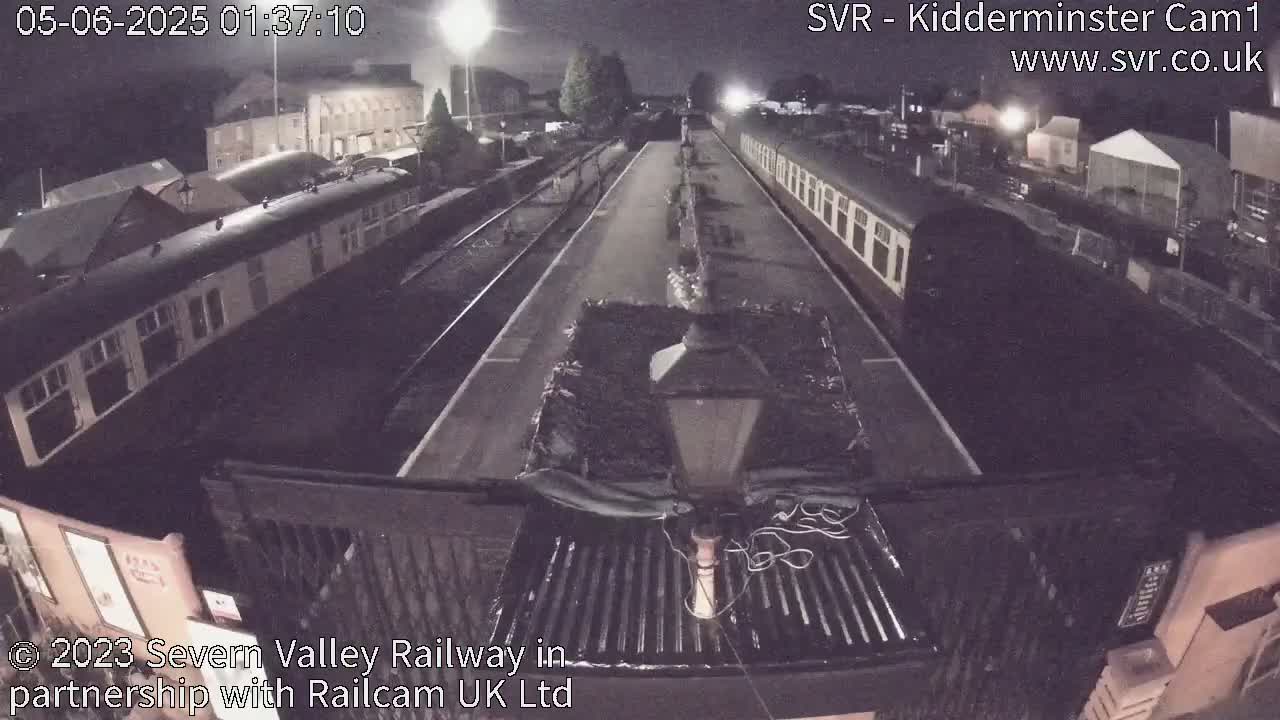 A nighttime, high-angle view shows a train station with railway cars parked on tracks under artificial lighting.