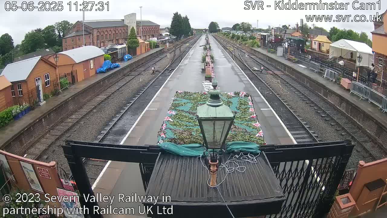 A wet train station platform with multiple train tracks stretches into the distance, featuring several small buildings and a camouflaged object in the center.