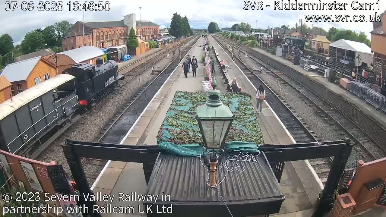 A train pulls into a railway station on an overcast day, with several people walking on the platform.