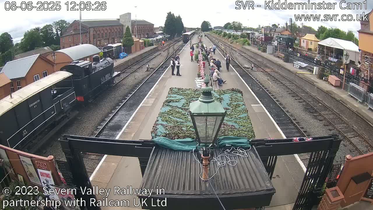 A black steam train is arriving at a railway station on a partly sunny day, where several people are standing and sitting on the platform.