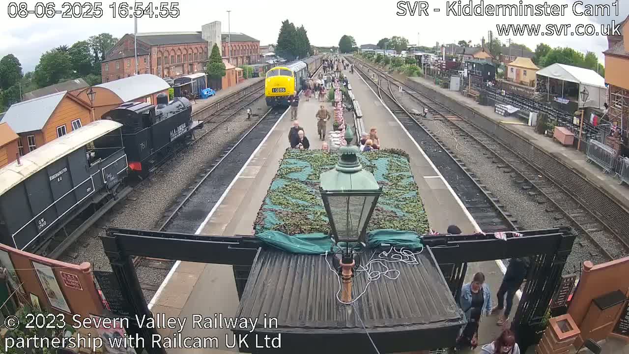 A railway station platform with several trains, people walking, and a camouflaged object on a train, under partly cloudy skies.