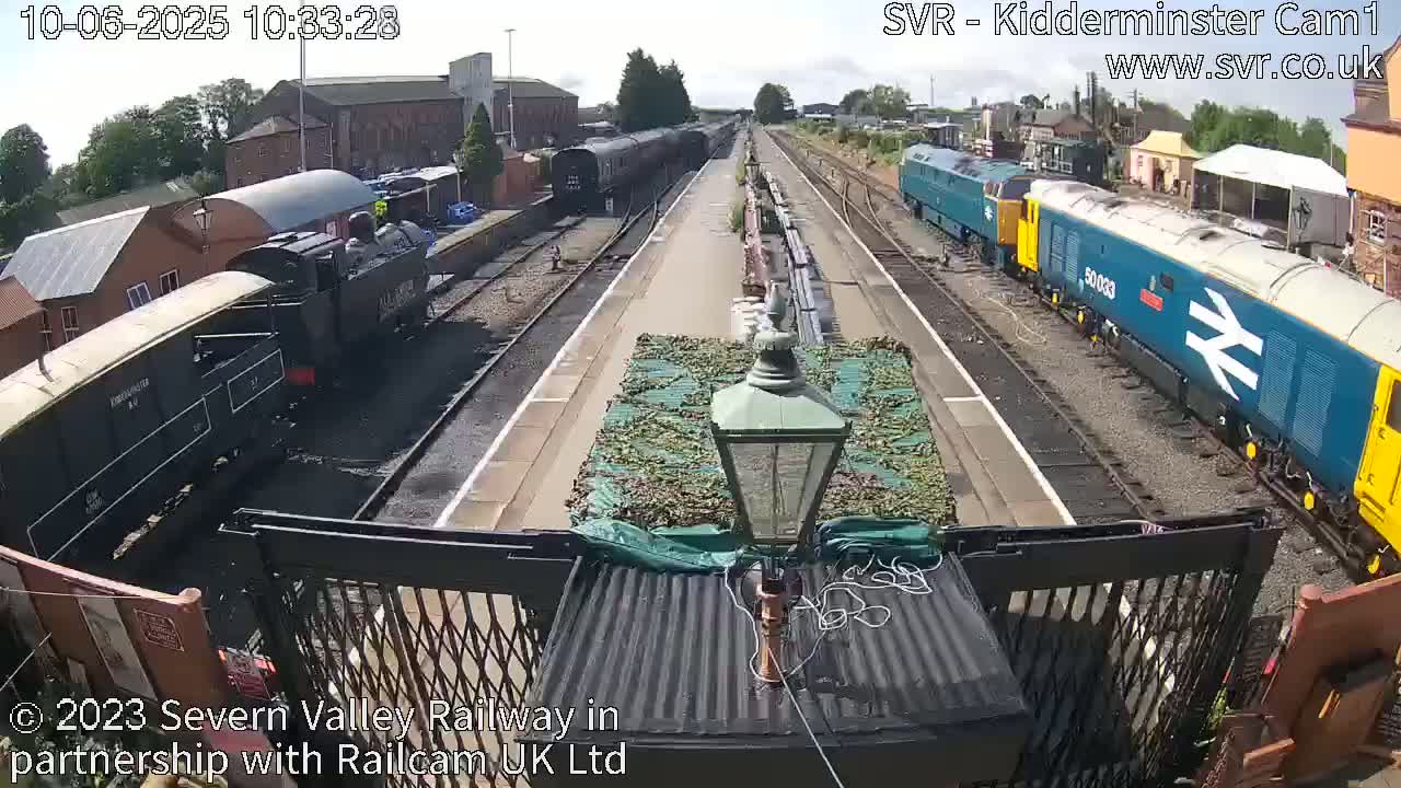 A sunny day at a train station shows multiple locomotives on the tracks, with one dark-colored steam locomotive in the foreground and a blue diesel locomotive further down the tracks.