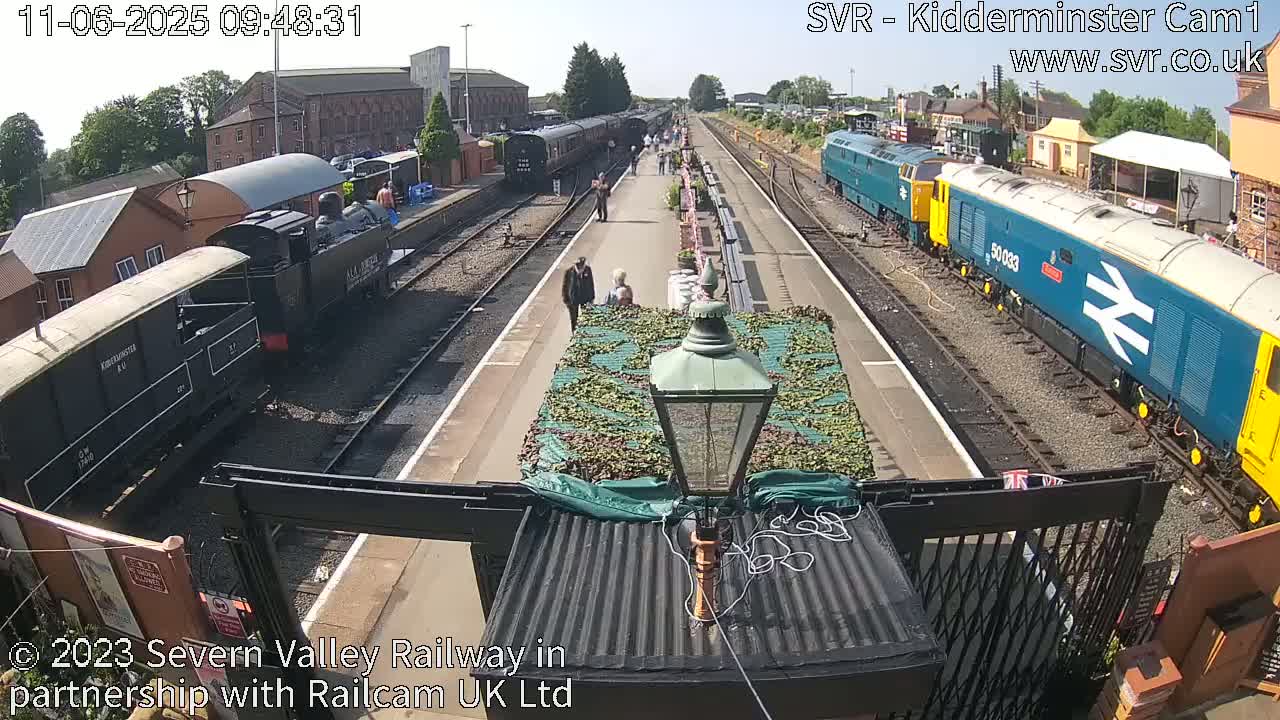 A sunny day at a train station shows several locomotives and passenger cars, some people walking on the platform, and a partially covered structure in the foreground.