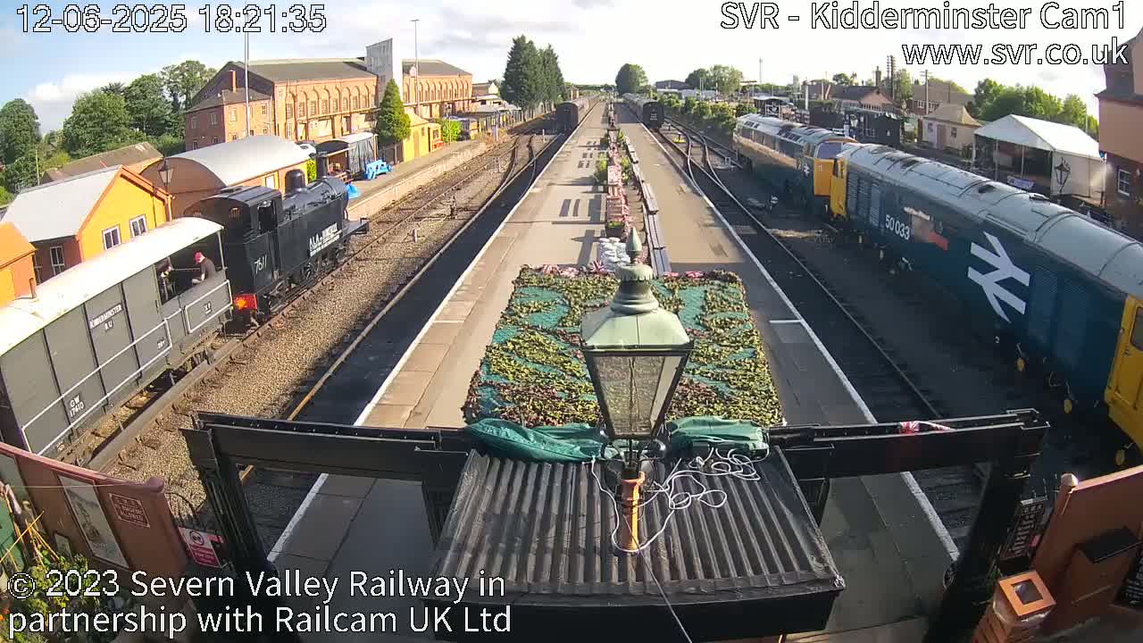 A sunny day at a train station shows several trains on tracks, one of which is arriving, with buildings and foliage in the background.
