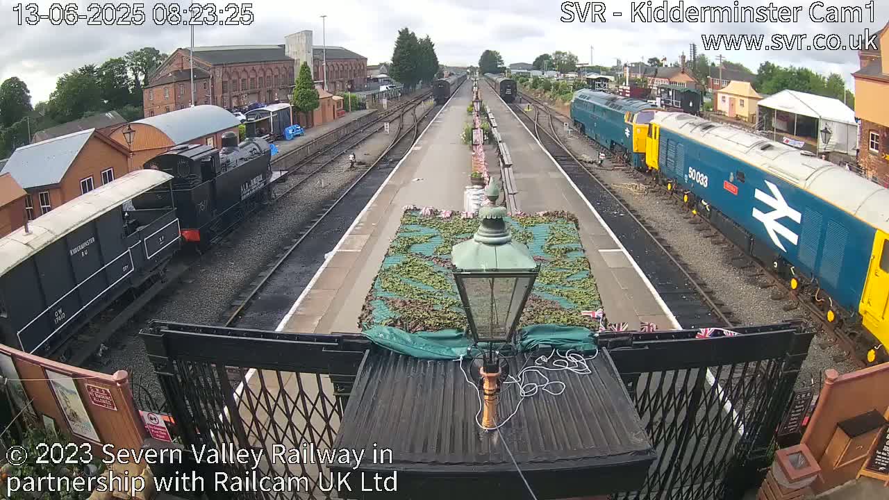 A railway station platform with several trains, including a dark-colored steam locomotive and a blue diesel locomotive, under a mostly cloudy sky.