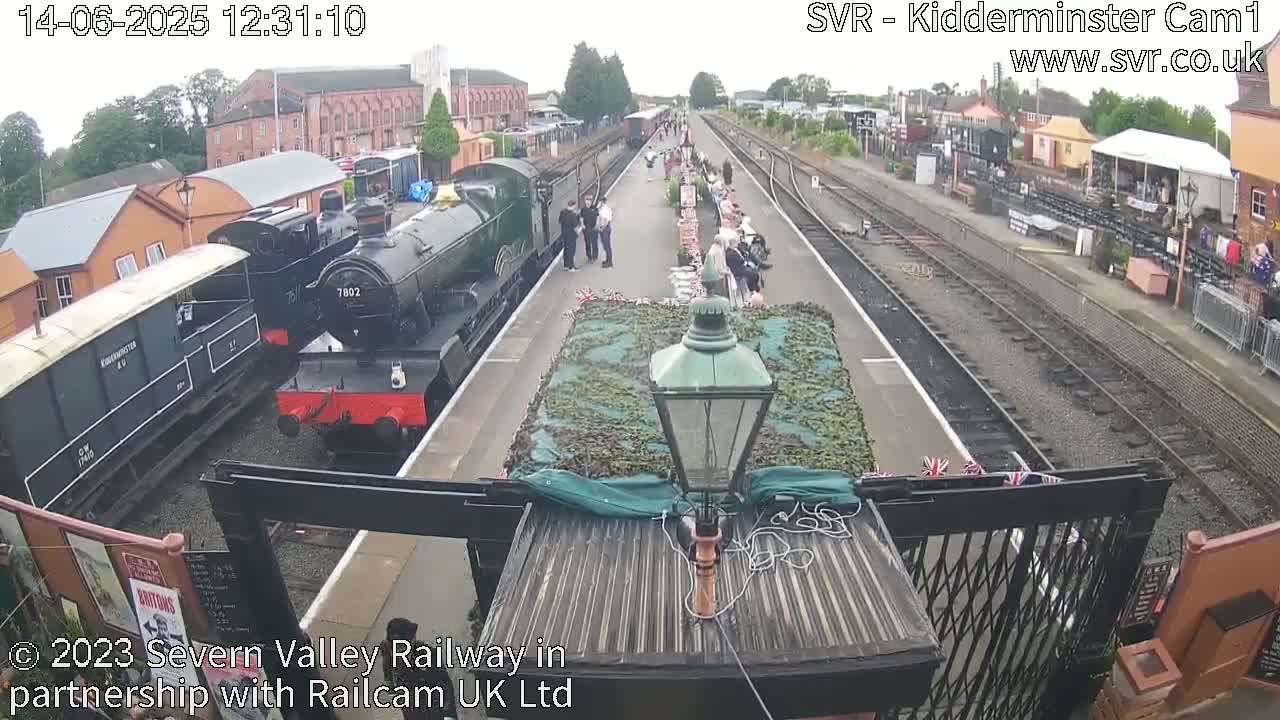 Several steam trains are parked at a railway station on an overcast day, with people gathered on the platform.