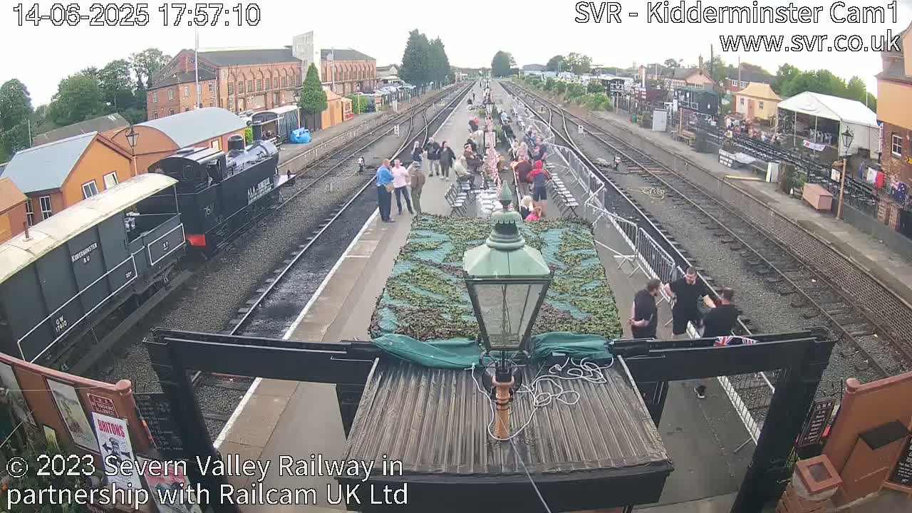A train pulls into a railway station where several people are gathered, under an overcast sky.