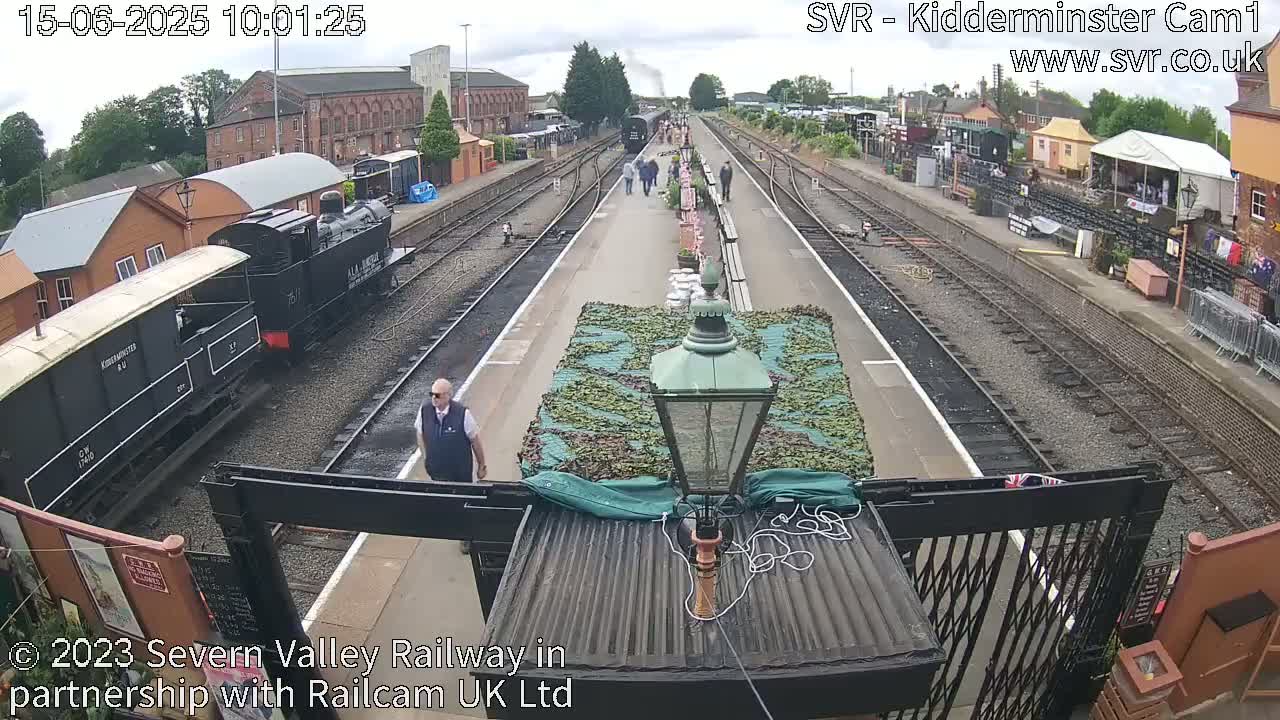 A train station platform on a partly cloudy day with a steam train and several people walking on the platform.