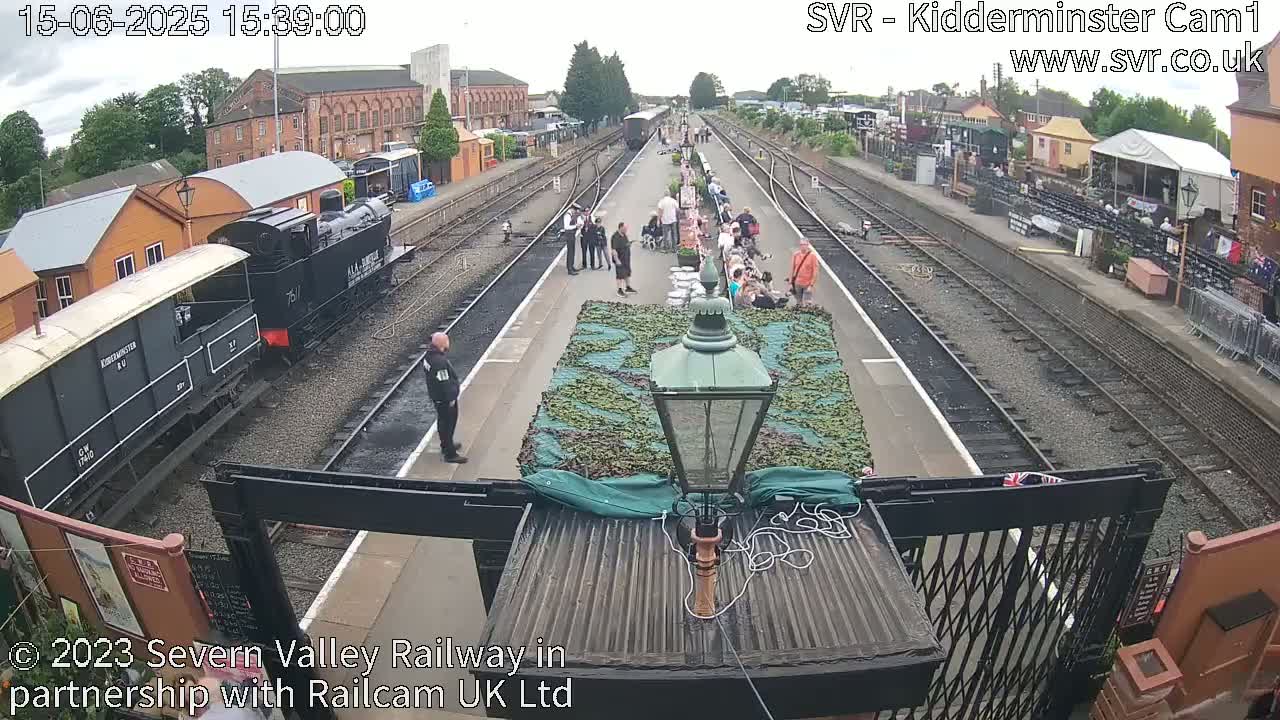 A railway station platform with several people gathered, a steam train partially visible, and a camouflage-covered object in the center, under a partly cloudy sky.