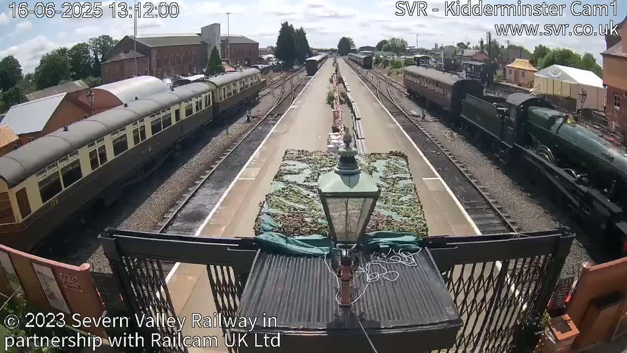 A sunny day at a train station shows two passenger trains and a steam locomotive on parallel tracks.