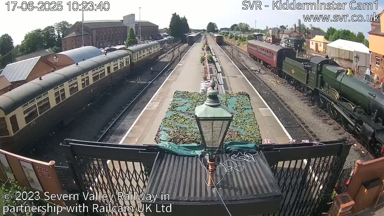 A sunny day at a train station shows two passenger trains on parallel tracks, one dark brown and one dark green, with a partially covered area in between them.