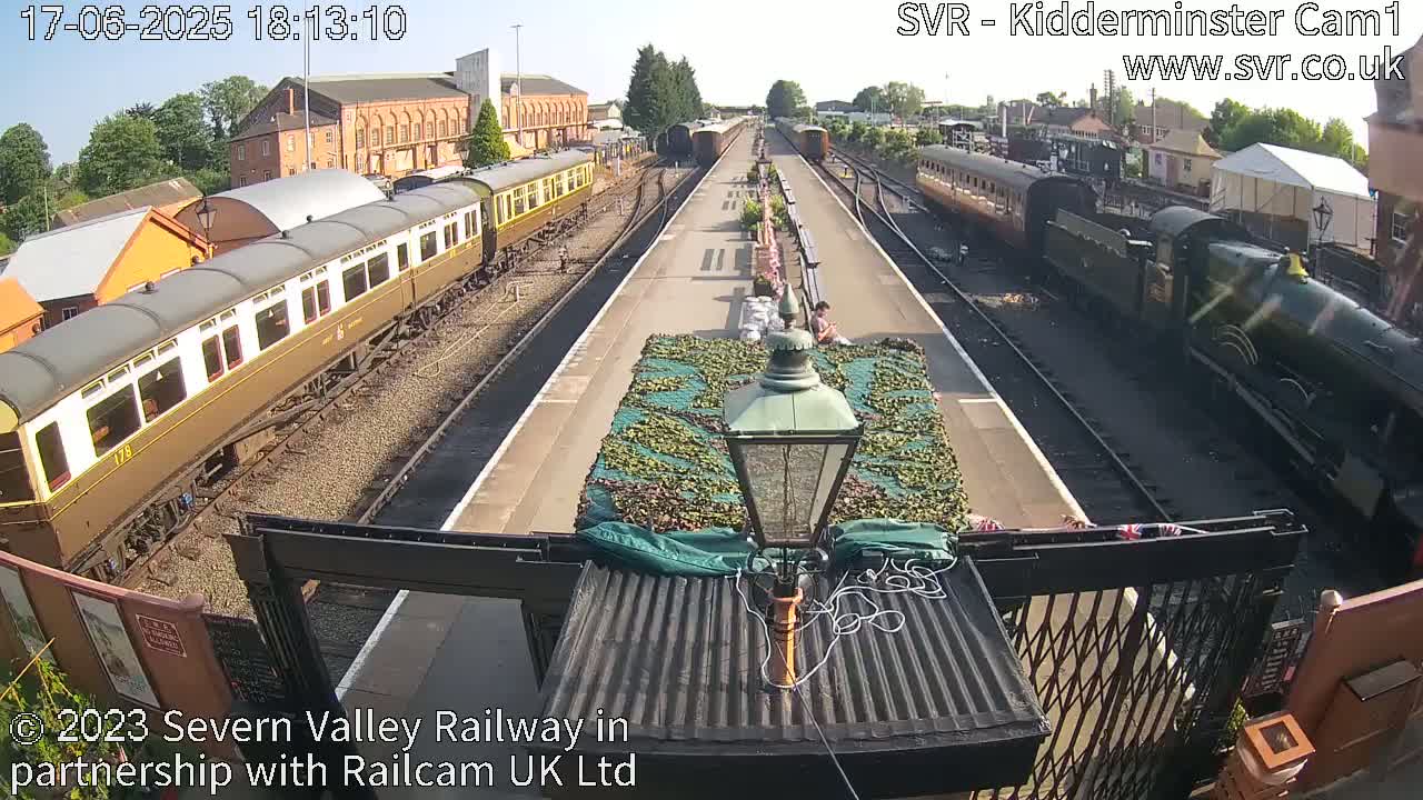 A sunny view of a railway station with passenger train cars on either side of a platform, a steam locomotive at one end, and a camouflage-covered object in the center.