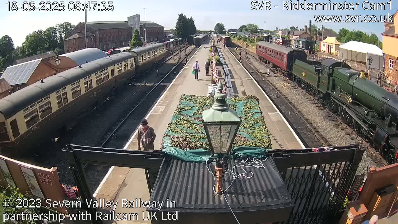 A sunny day at a train station shows several passenger cars and a steam locomotive on parallel tracks, with a few people walking on the platform between them.