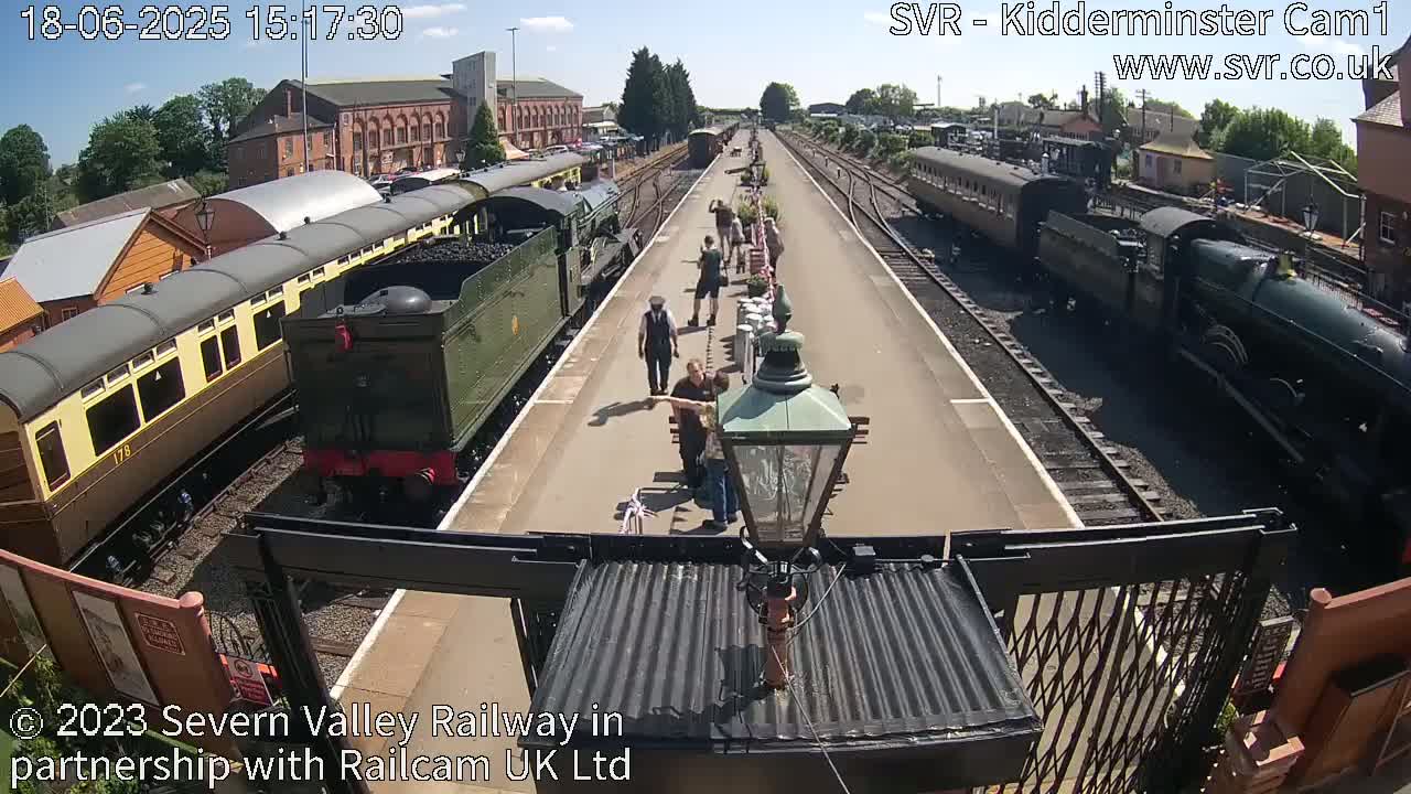 A sunny day at a train station shows several vintage passenger cars and locomotives on the tracks, with several people standing on the platform between them.