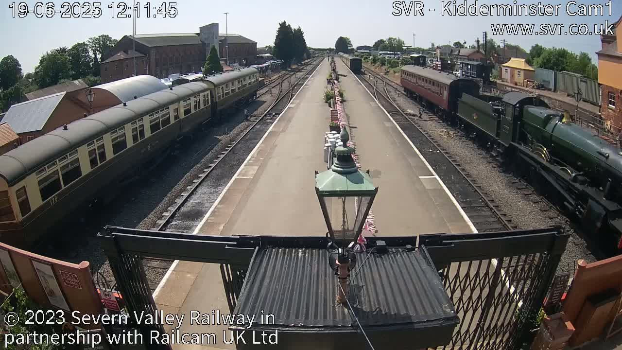 A sunny day at a train station shows a long platform with passenger cars and a steam locomotive on either side.