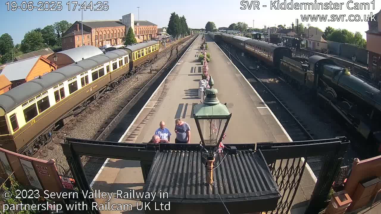 A sunny day at a train station shows two passenger trains and several people standing on the platform.