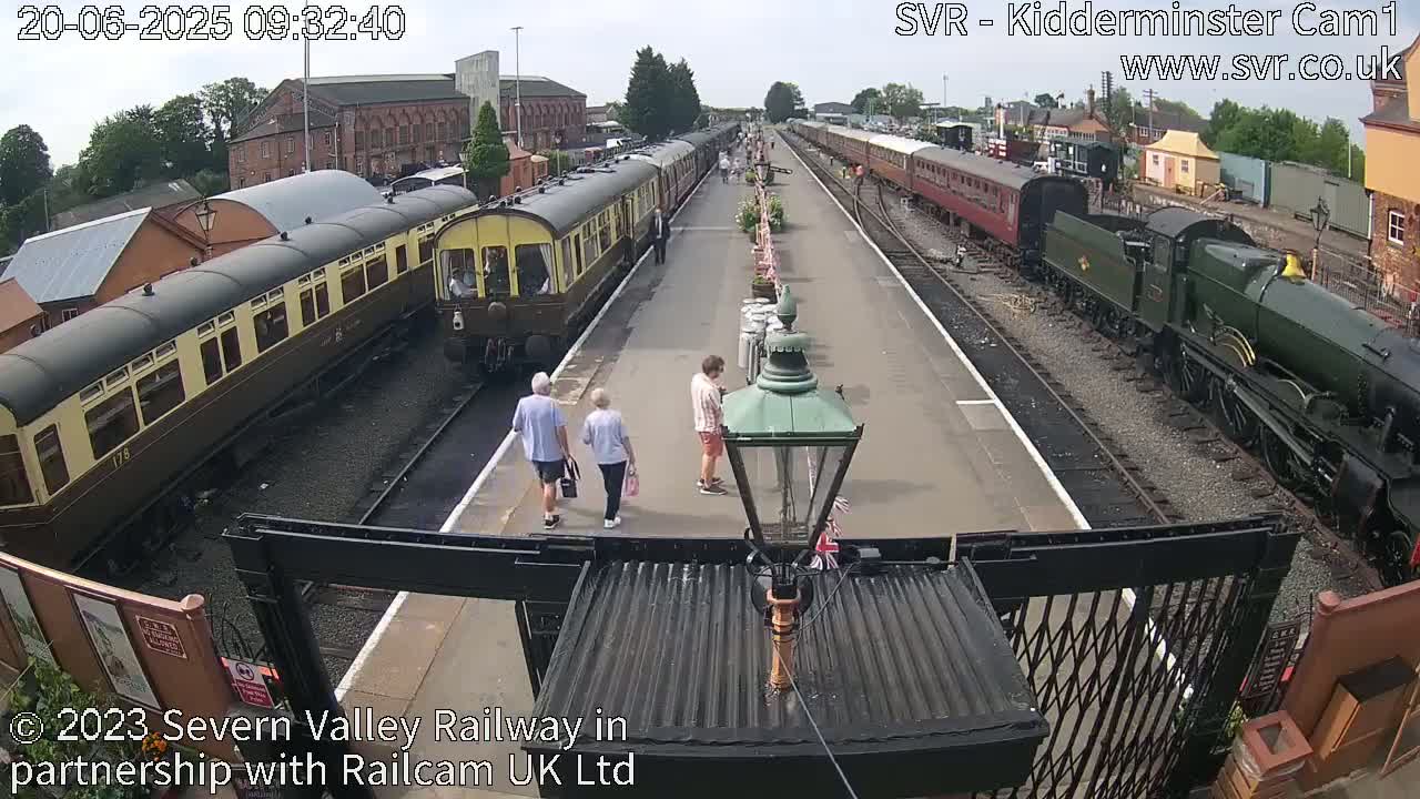 A sunny day at a train station shows several passenger cars and locomotives on the tracks, with a few people walking on the platform.