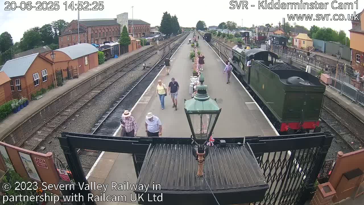 A dark-green steam train sits at a railway station platform where several people stroll on a partly sunny day.