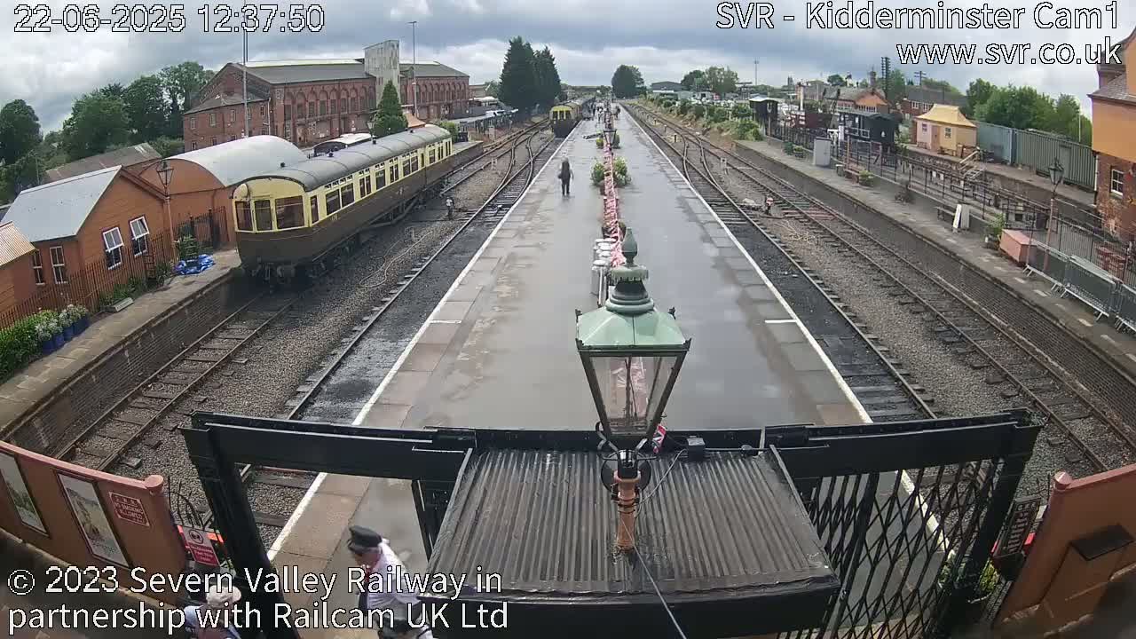 A railway station platform with a train and several people, under an overcast sky.