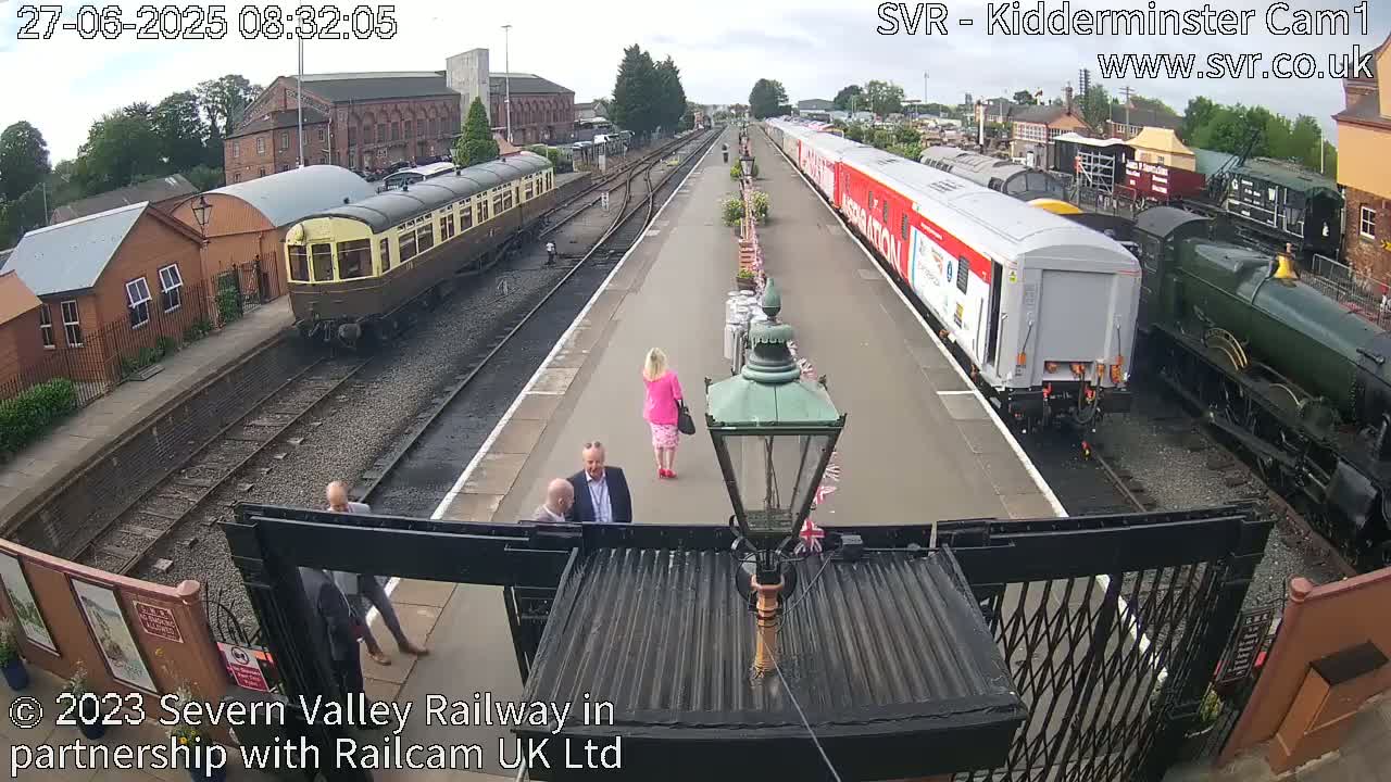 A sunny day at a train station shows several passenger trains and a steam locomotive parked on the tracks, with a few people walking on the platform.