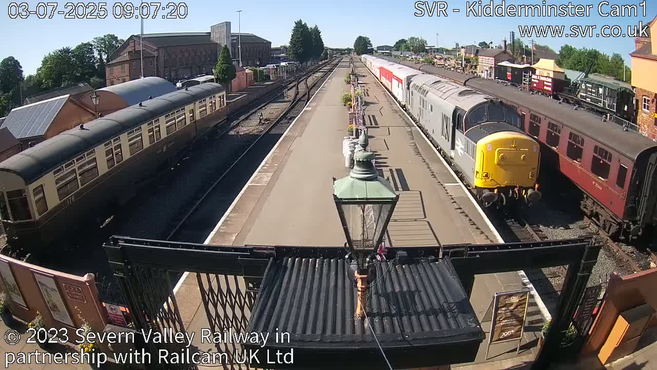 A sunny day at a train station shows several passenger train cars and locomotives on parallel tracks, with one train departing.