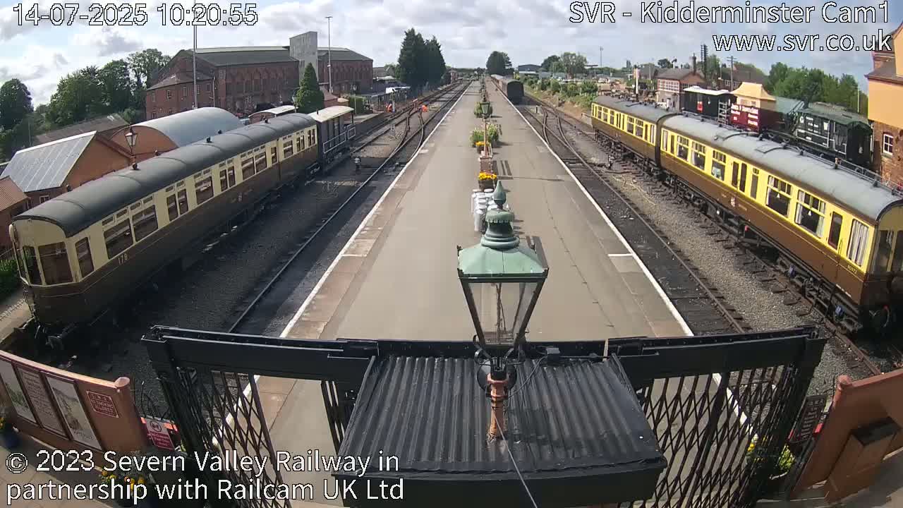 Two long passenger train cars are parked on parallel tracks at a railway station on a partly sunny day.