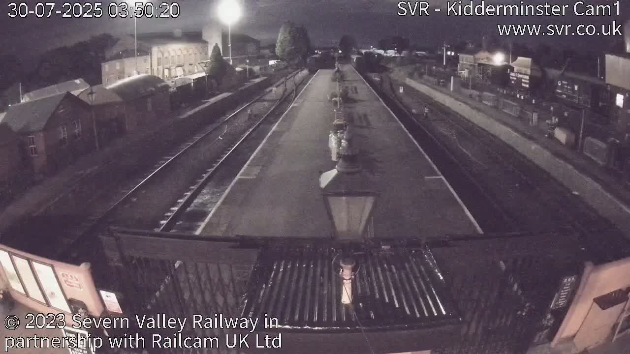 A wet train station platform with a long passenger train parked alongside it, seen from behind a gate under a gray sky.