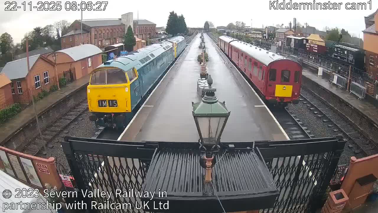 An overhead view reveals two trains – one blue and yellow, the other red – stopped at a wet platform in a train station, under an overcast sky.