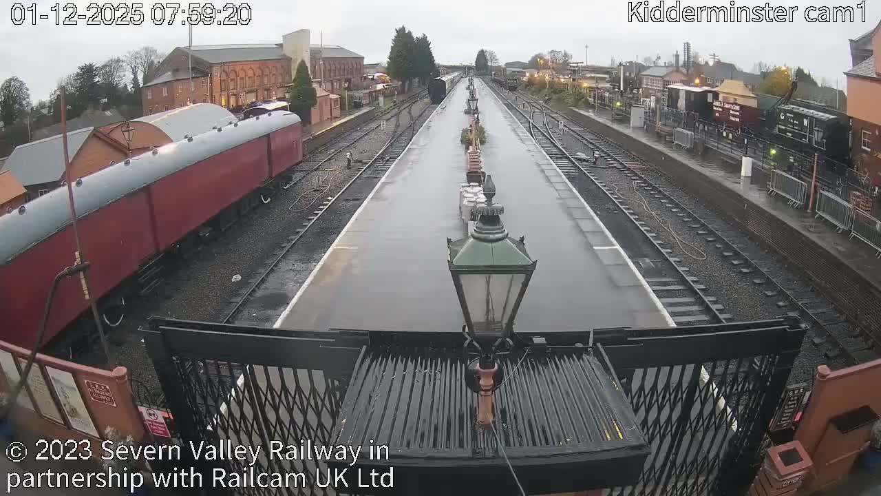 A wide view of a classic railway station displays wet platforms and numerous tracks, with a long red passenger carriage on the left and a dark freight train on the right, all under an overcast and damp sky.