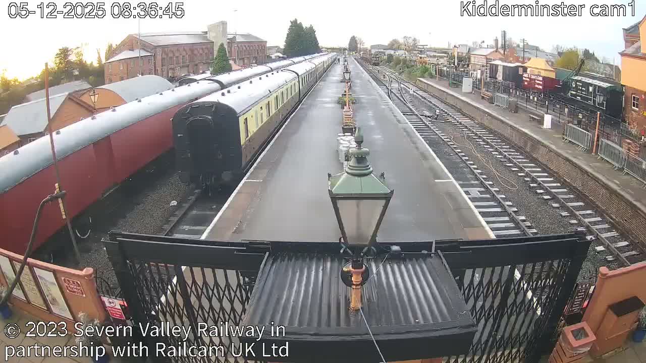 An elevated view captures a train station with a long, empty platform lined with lamps, a vintage train parked to the left, and additional tracks and freight cars to the right, all under a bright, clear sky.